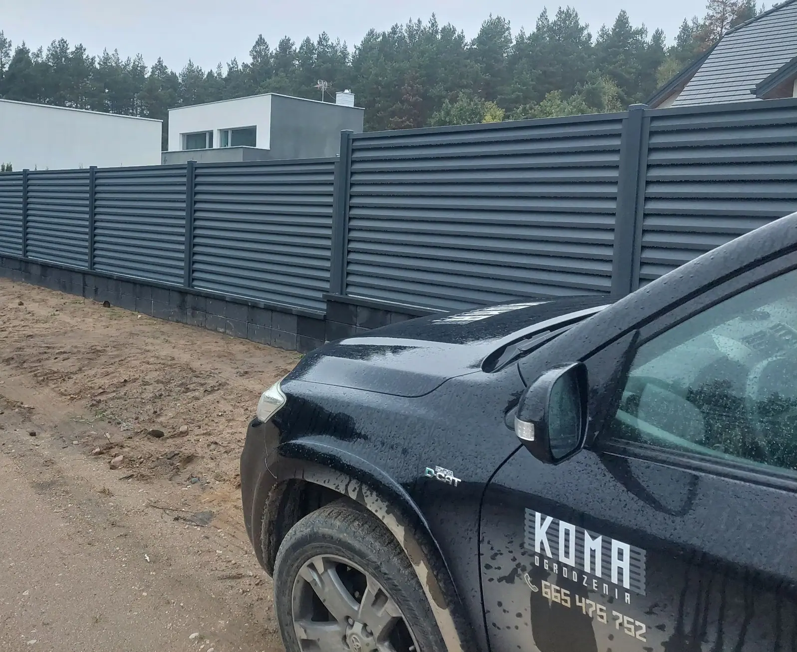 Samochód firmy KOMA z logo na drzwiach przy ogrodzeniu żaluzjowym w kolorze antracytu